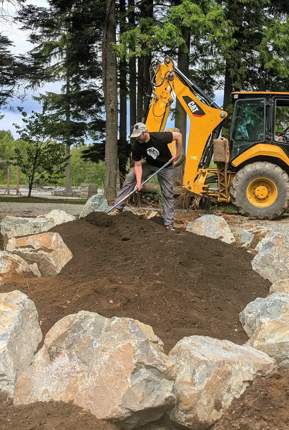 A backhoe filling a ring of boulders with soil beside the cedar building