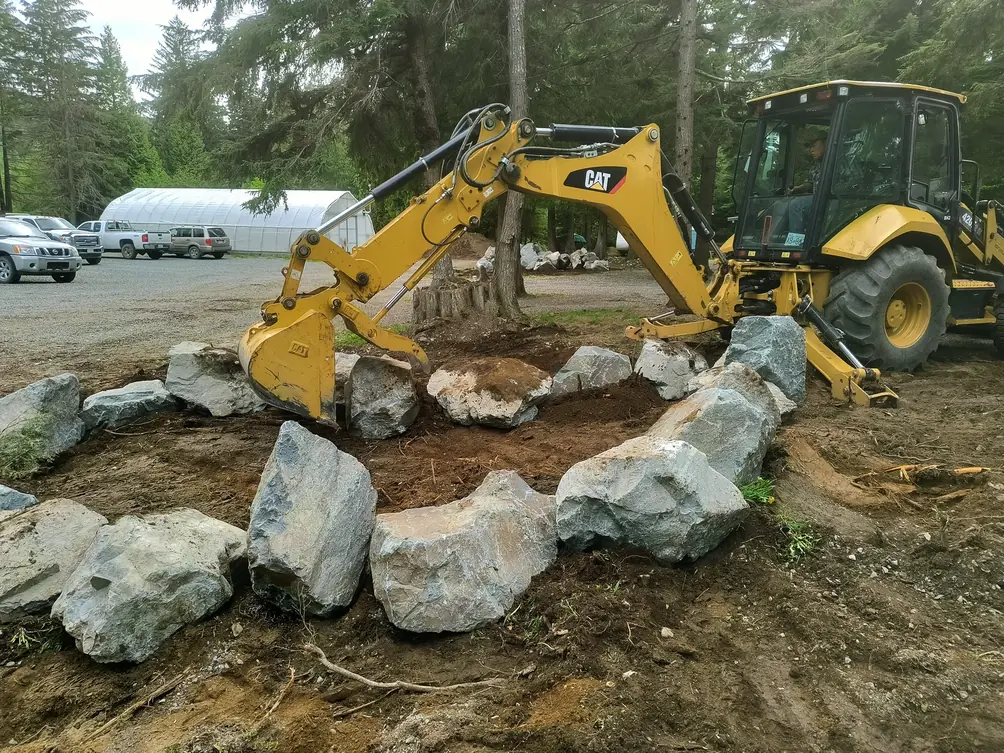 A loader working soil inside a ring of boulders among the cedars